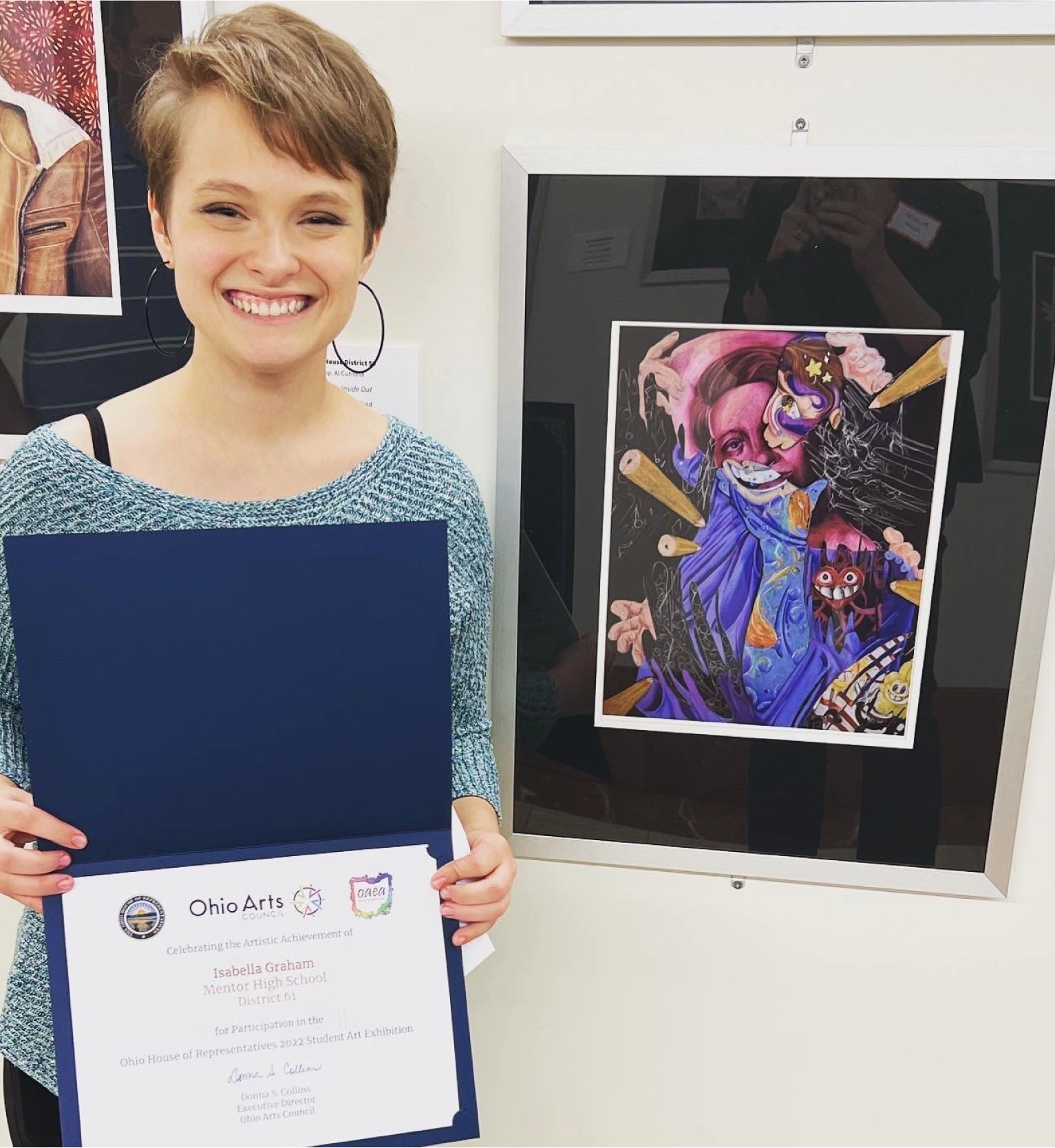 Photograph of student posing next to artwork. Student is holding a certificate of achievement from Ohio Arts.