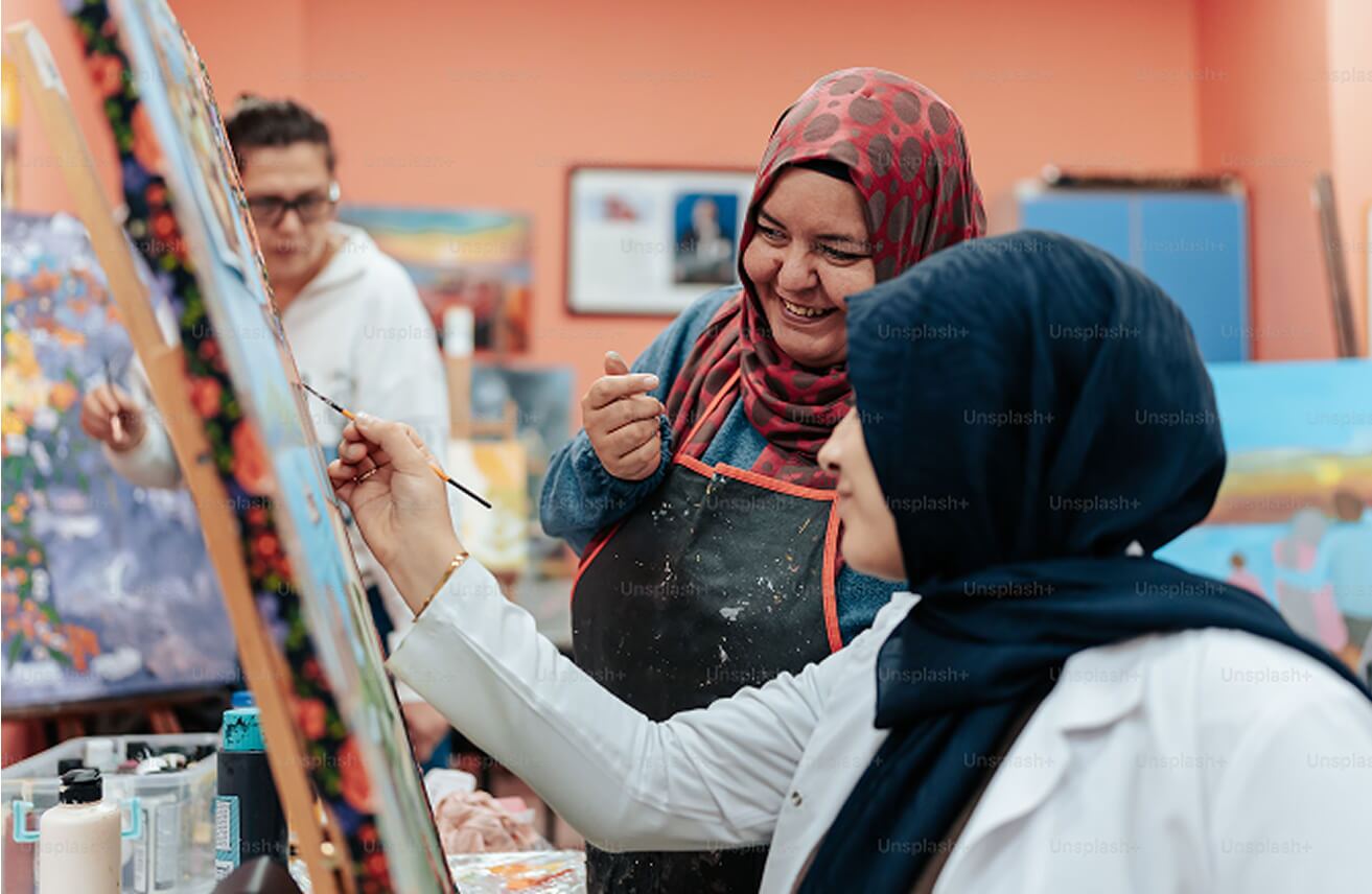 two women wearing head coverings. one of them is working on a painting in progress, and the other is watching her work while smiling