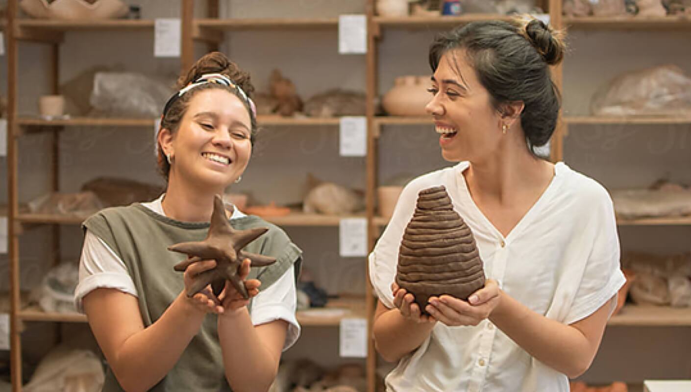two women holding up clay sculptures and smiling while looking at each other