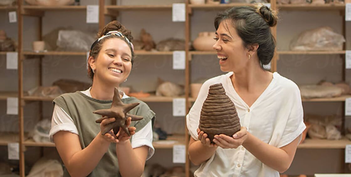 two women holding up clay sculptures and smiling while looking at each other