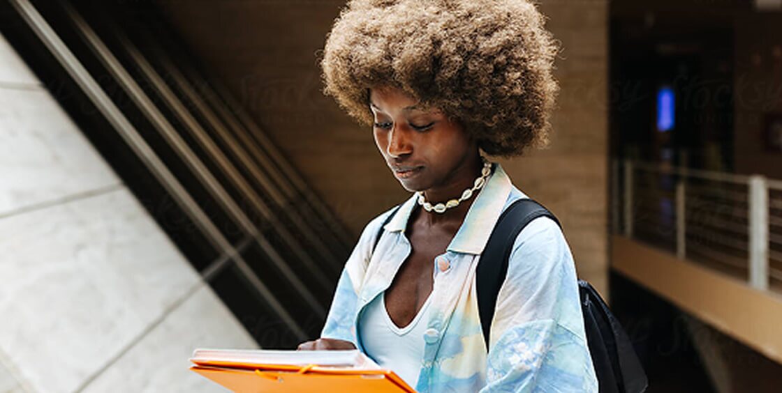 young woman wearing a backpack and holding a stack of books and orange folders. She appears to be examining the book