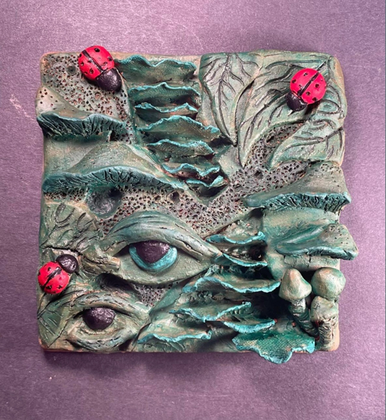 Tile-sized sculpture depicting leaves, mushrooms, eyes, and ladybugs