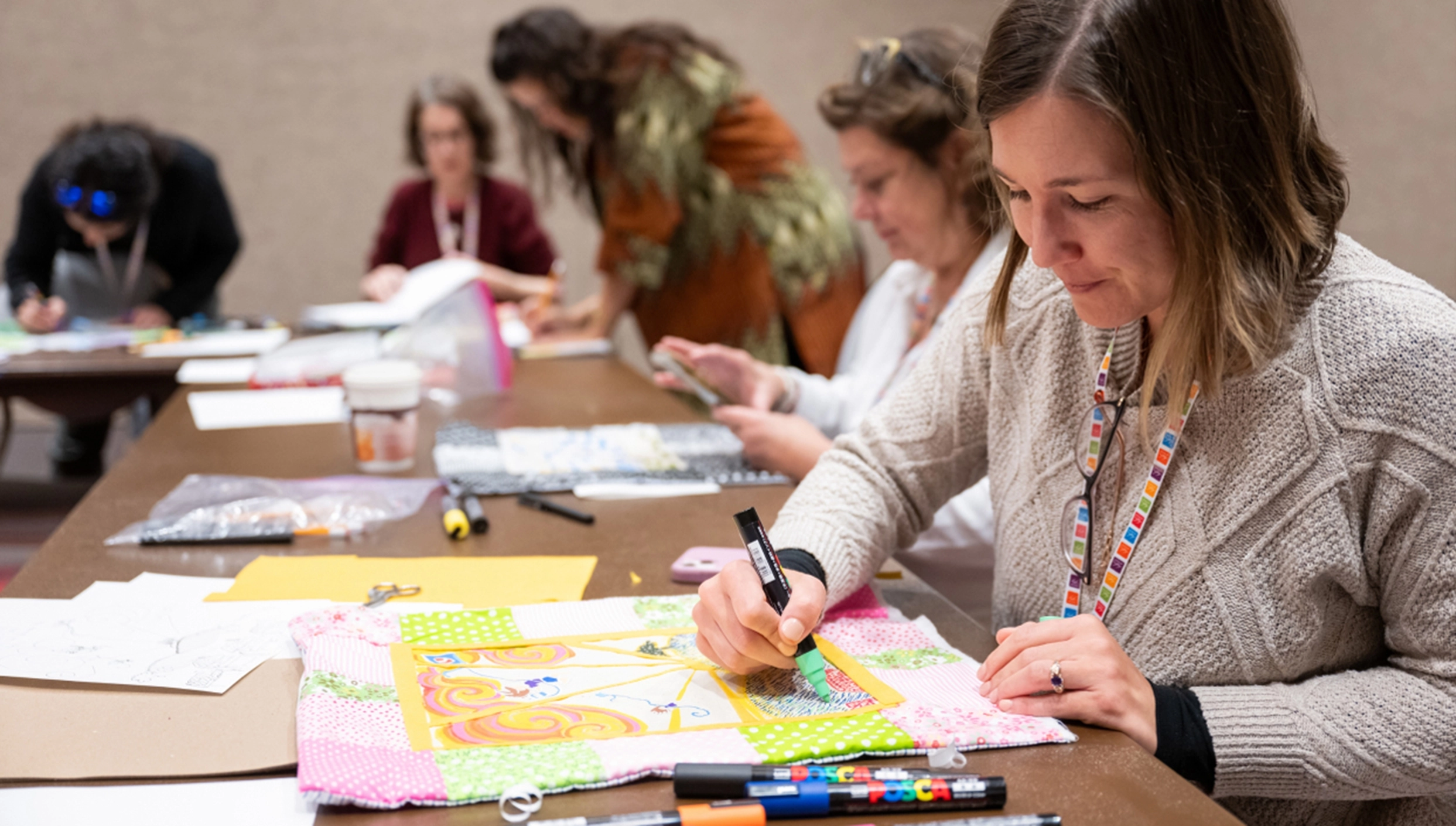 people seated at long tables drawing on quilt squares with Posca paint markers